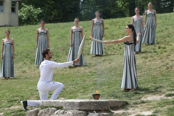 扮演最高女祭司的演員瑪麗·米娜（前右）點燃第一棒火炬手——希臘運動員、東京奧運會男子賽艇項目金牌得主斯特凡諾斯·恩圖斯科斯手中的火炬。新華社記者 趙丁喆 攝